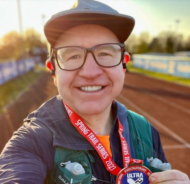 Ben smiles at the camera with his first medal for completing a 5km ultra. He wears a cap and glasses