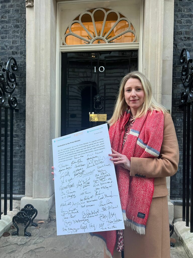 Sarah Quinlan MBE in front of 10 Downing street delivering a petition. She wears a fawn coat and a big pink scarf and had fair hair. Her expression is serious.