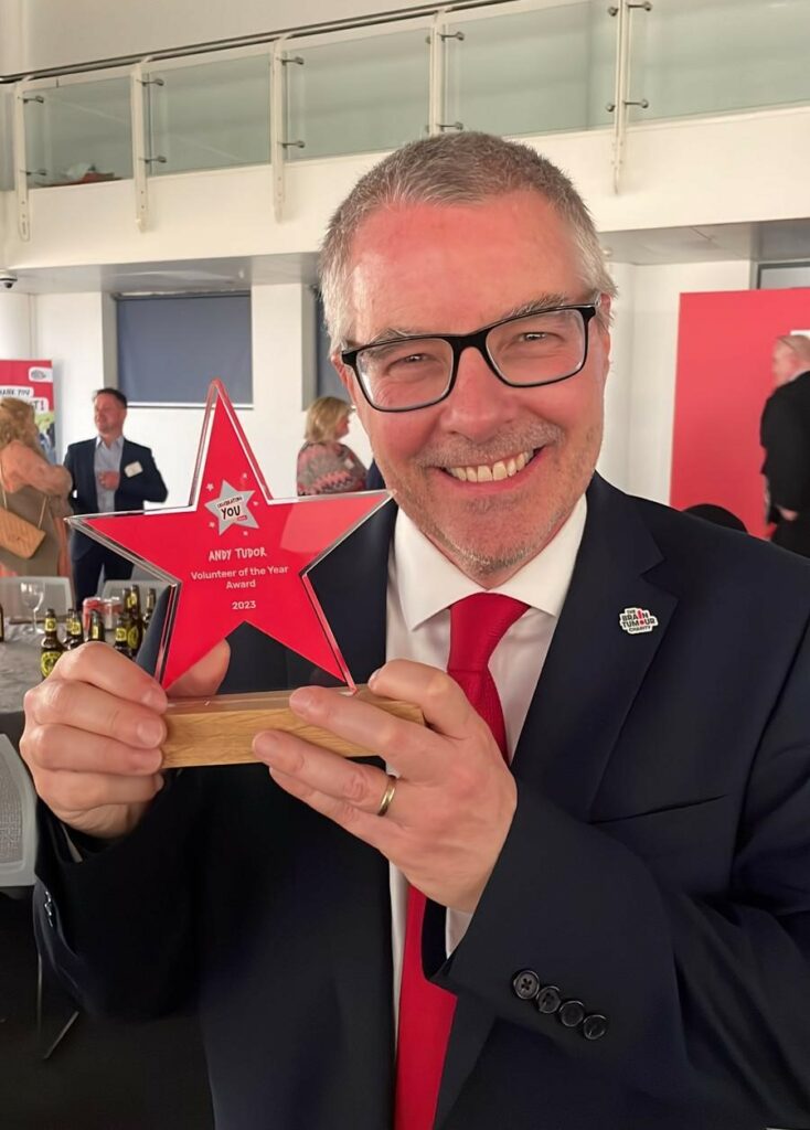 Andy Tudor holds his volunteer award and smiles at the camera. He has short hair, black glasses and a smart jacket with a red tie
