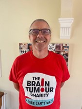 Andy Tudor has short hair and glasses and beams at the camera, wearing The Brain Tumour Charity t-shirt which is bright orange