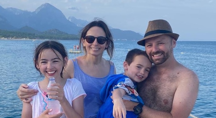 Chris Ruggles pictured on holiday with his wife, Jenny and their two children. Everyone is smiling and there is water and mountains in the background. Chris wears a hat and Jenny wears sunglasses