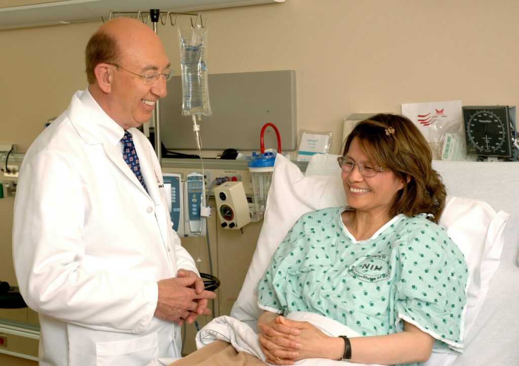 A woman with dark hair and olive skin wears a hospital gown and sits in a hospital bed. She is talking with a male doctor and there is medical equipment in the background