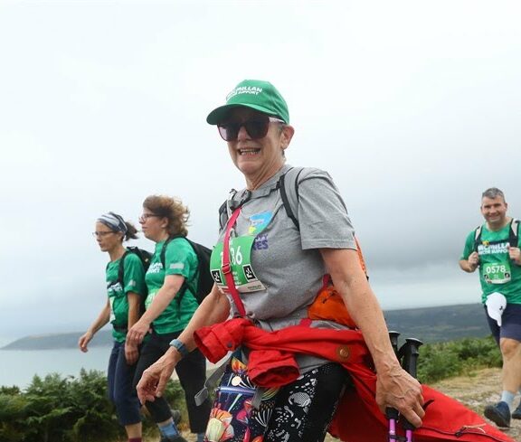 Dee hikes with friends, smiling at the camera. She wears sunglasses and a green cap and ha short hair. Her t-shirt is grey and a bright red hoodie is tied around her waste