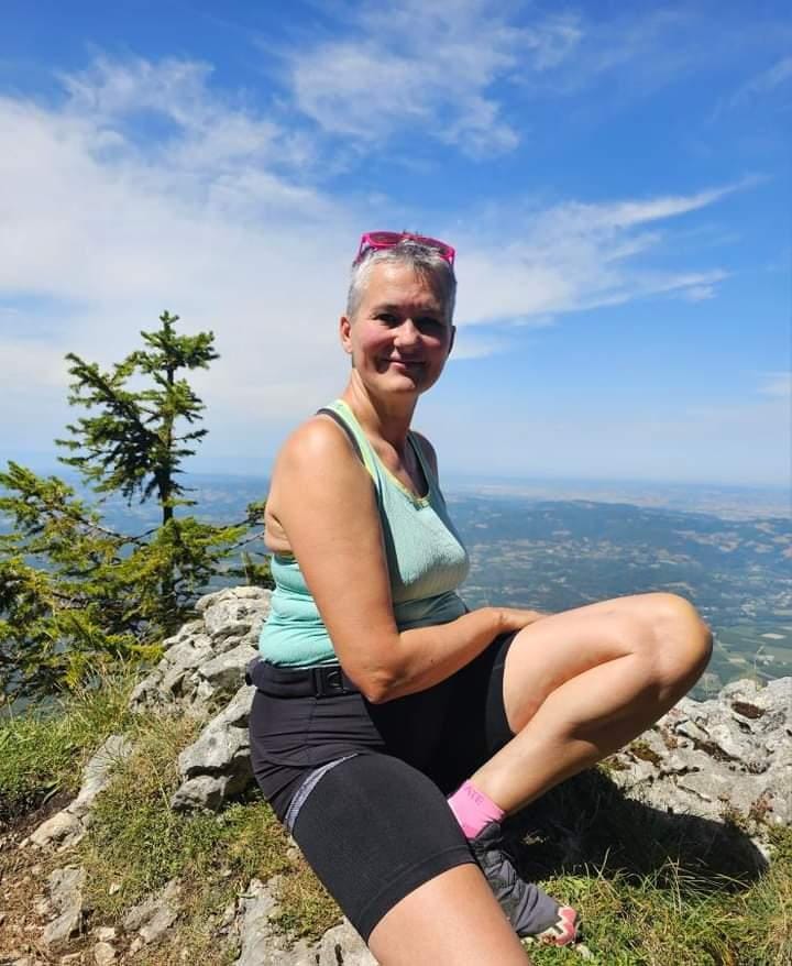 Georgi sits on a rock, on a hillside, in front of a rbight blue sky. She is smiling, her hair is short and she wears a green tshirt and black shorts.