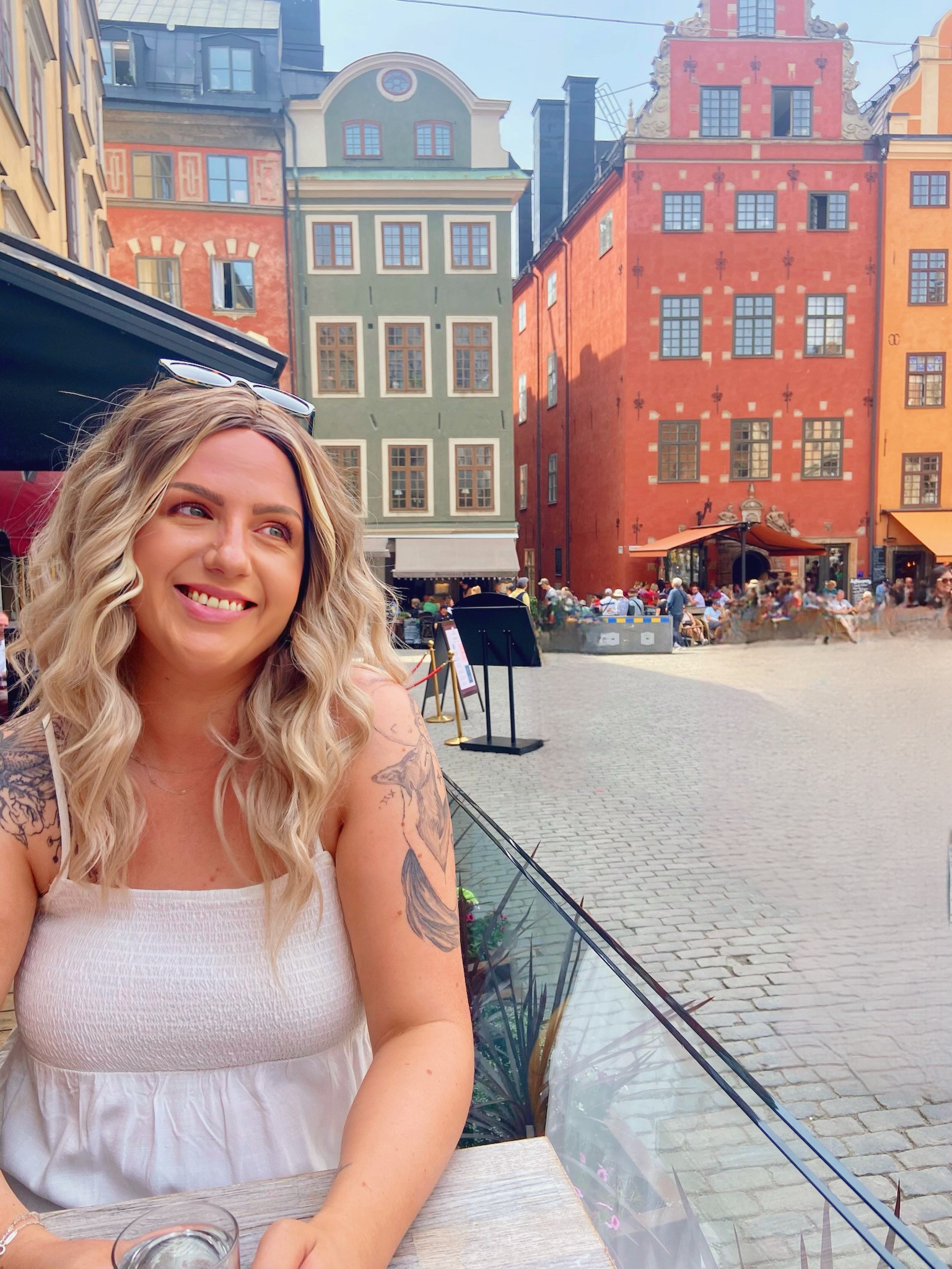 Jessica in Stockholm, on a holiday with her friend. She smiles, wears sunglasses on her head and has a white summer dress and long wavy fair hair