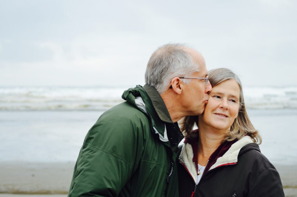 An older man and his wife stan close together on a beach in winter. They wear warm coats and the man is kissing the woman on the cheek. He wears glasses and she smiles