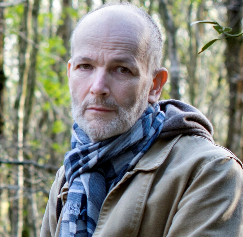 marcus has trees behind him. He has short hair, a grey jacket and a blue checked scarf. He has a white beard and a serious expression