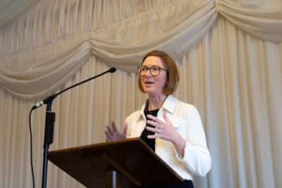 Dr Katie Wakeham speaking at the launch of a 10 year Vision for Radiotherapy. She wears a white jacket and has bobbed, brunette hair and glasses, speaks into a microphone and gestures with her hands 