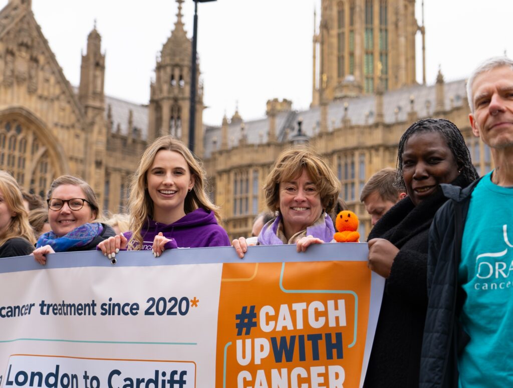 Supporters of the Catch Up With Cancer campaign hold an orange and cream banner in front of Westminster. They are smiling at the camera.