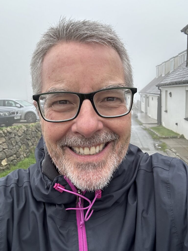 Andy smiles at the camera. He has short grey hair and grey facial hair and wears black glasses. He is walking in the countryside on a wet day