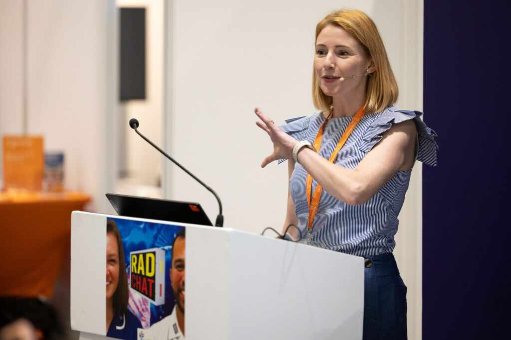 Sarah Quinlan wears a blue top and skirt and speaks from behind a podium. Her hair is strawberry blonde and in a bob and she gestures with her hand.