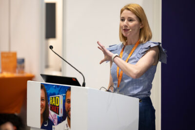 Sarah Quinlan wears a blue top and skirt and speaks from behind a podium. Her hair is strawberry blonde and in a bob and she gestures with her hand. 