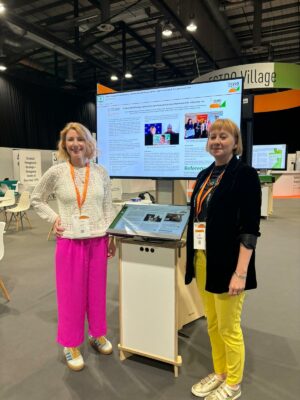 Sarah and Lynsey from Radiotherapy UK stand next to our abstract poster, shown on a digital screen