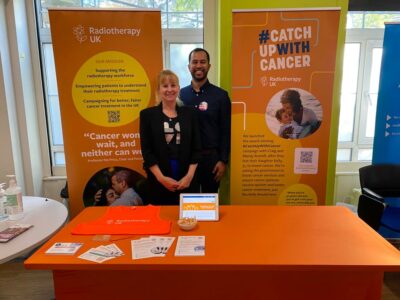 Lynsey Rice (Left) and Ambassador Naman Julka-Anderson stand in front of two bright orange banners at a conference for oncology professionals