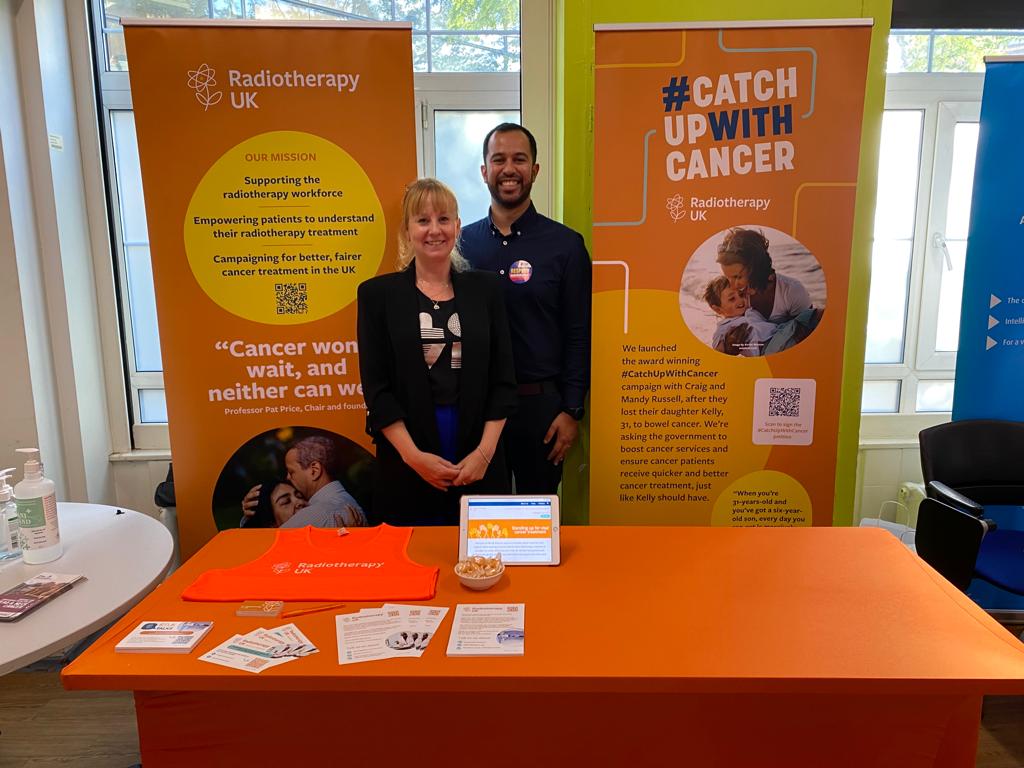 Lynsey Rice (Left) and Ambassador Naman Julka-Anderson stand in front of two bright orange banners at a conference for oncology professionals