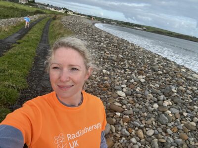 Sarah Quinlan MBE, wears her orange Miles 4 Radiotherapy t-shirt on a pebble beach. She has blonde hair and the weather is cloudy