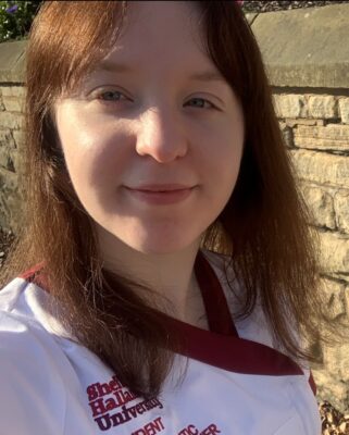 Emily Reed has brown shoulder-length hair and a fringe. She has pale skin and smiles at the camera on a sunny day