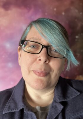 Dr Char Goodwin has blue hair swept to the side and wears black glasses. She has a friendly, thoughful expression and looks straight into the camera