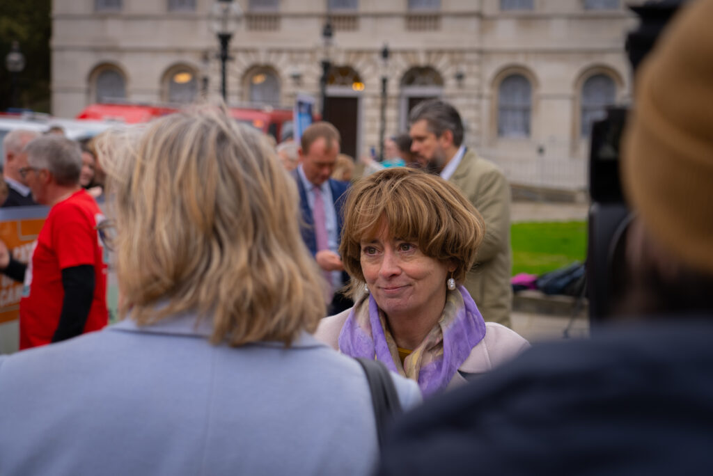 Pat Price speaks to the media outside Westminster. She has a brown bob, purple scarf and a cream jacket