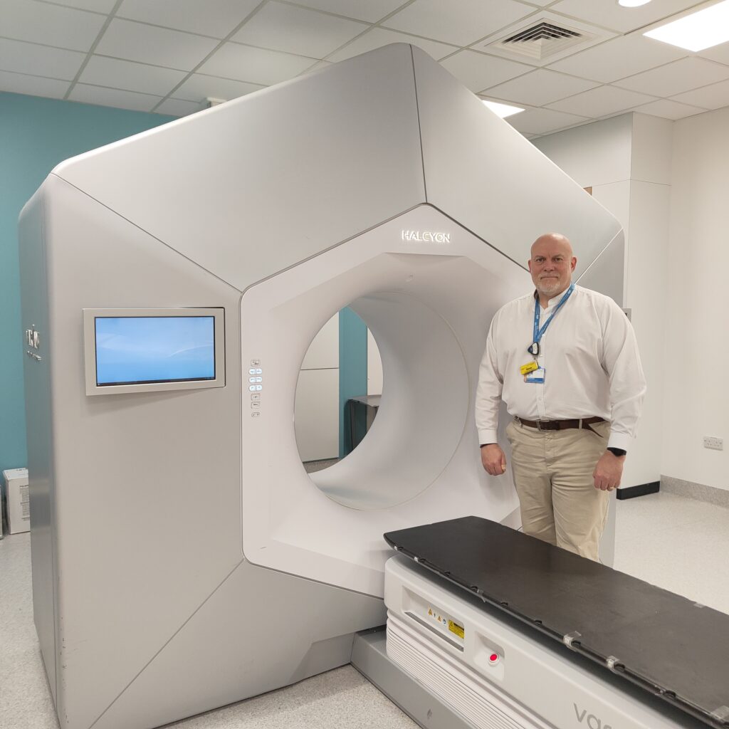 Dom Withers wears a white shirt, beige trousers and a blue lanyard. He stands in front of a Linear Accelerator, which is a radiotherapy treatment machine