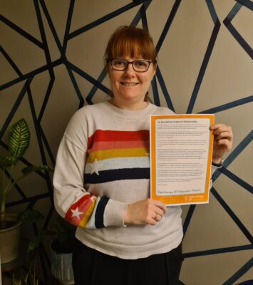 Emma from RTUK has brown hair and glasses and holds the letter written by Ambassadors. She wears a colourful jumper and is smiling. 