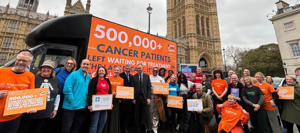 Campaigners crowd around a huge digital van outside Westminster, which reads: 500,000+ Cancer Patients left waiting for treatment in the last decade. It’s time to #CatchUpWithCancer. Change.org/catchupwithcancer. The campaigners hold orange signs and wear bright clothes