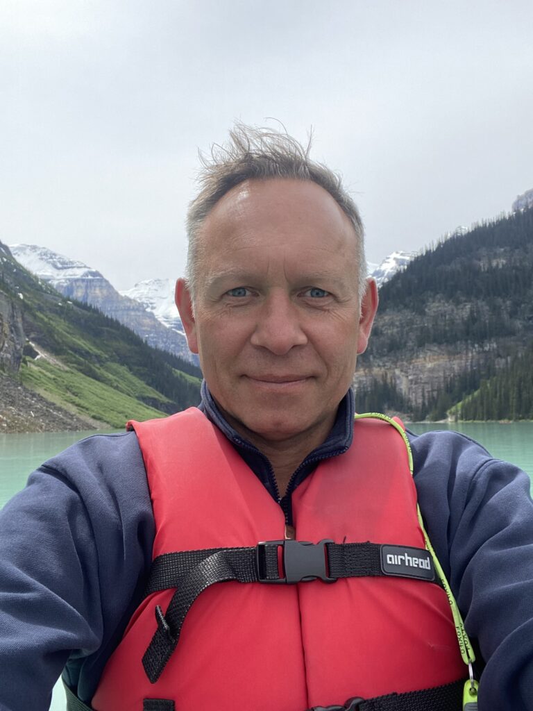 Jeff Gibson has fair hair and tanned skin. He wears a life jacket and seems to be sailing on a lake with snowy mountains in the background