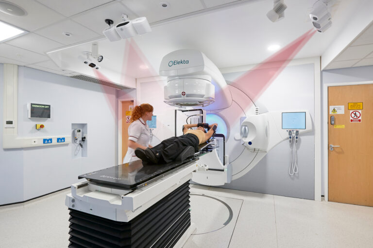 A radiotherapy professional positions a patient carefully on a radiotherapy machine. This is like a horizontal bed for the patient to lie on while a large structure moves around them, delivering a careful dose of radiation