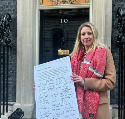 Sarah Quinlan MBE in front of 10 Downing street delivering a petition. She wears a fawn coat and a big pink scarf and had fair hair. Her expression is serious.